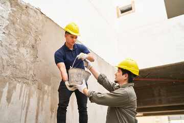 Young male coworkers in safety workwear carrying bucket by wall at construction site