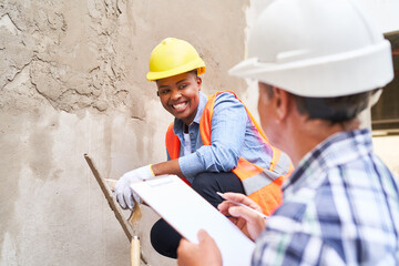 Happy female bricklayer with trowel crouching near senior builder at construction site