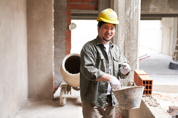Portrait of smiling young male bricklayer standing with bucket by incomplete wall in corridor at...