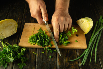 Slicing fresh parsley with a knife in the hands of a cook on a cutting board. Low key concept of...