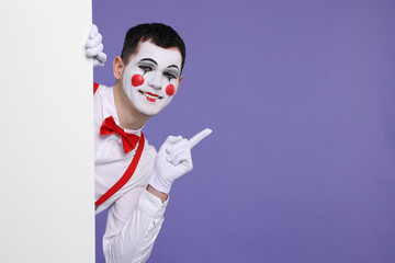 Funny mime artist peeking out of blank poster and pointing at something on purple background. Space...