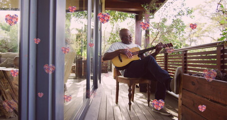 Image of pink flowers over senior african american man playing guitar on terrace at home - Powered by Adobe
