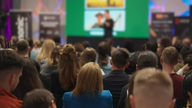Young audience engaged at entrepreneurship conference, female speaker presents on stage. Diverse group of men and women focused on business incubator event. Concept: entrepreneurship and startup