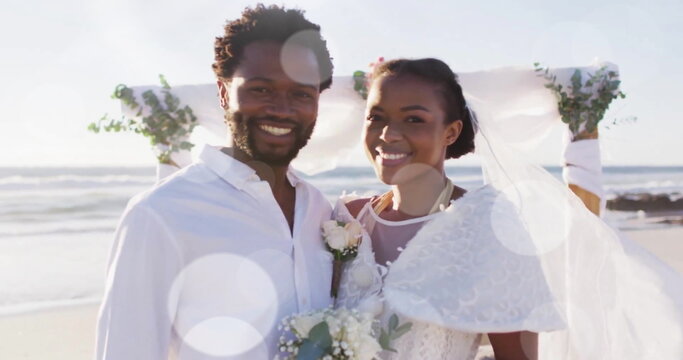Image of light spots over happy african american bride and groom smiling on beach at wedding