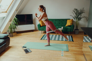 A woman in sportswear is performing exercises on a yoga mat in a living room with wooden flooring