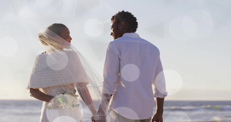 Fototapeta premium Image of light spots over african american bride and groom holding hands on beach at wedding
