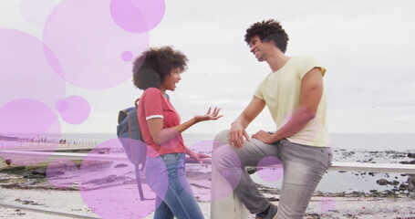 Fototapeta premium Image of purple spots over happy biracial couple talking on beach promenade