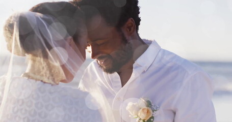 Naklejka premium Image of light spots over happy african american groom unveiling bride on beach at wedding