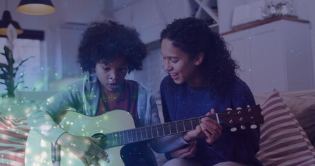Image of glowing spots over happy biracial mother with son playing guitar