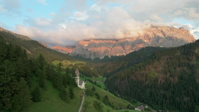 Aerial evening footage of the village of La Val with majestic Sas dles Nü (Cima Nove) mountain covered in clouds in the background. Drone flies close to church. South Tyrol, Italy. LuPa Creative