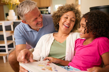 Multi Racial Grandparents Looking After Granddaughter At Home Reading A Story Together - obrazy, fototapety, plakaty