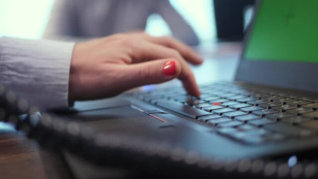 A female hand types a password on a laptop keyboard, dictated to her. The laptop screen has a green background for easy replacement with your content. The scene takes place in an office.