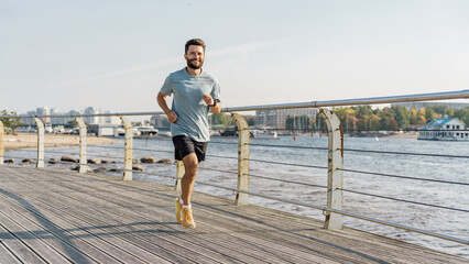 A joyful jogger with a light beard advances along a wooden pier by the river, cityscape and boats...