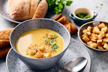 Bowl of mashed potato soup with a spoon on the plate. The bowl is filled with yellow soup puree and topped with croutons, parsley and ground red pepper.