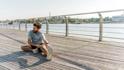 A bearded man engages in a seated leg stretch on a riverfront boardwalk, boats moored in the harbor...