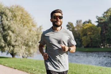 A runner with a beard and sunglasses jogs along a lakeside path, exuding confidence under the bright, open sky.