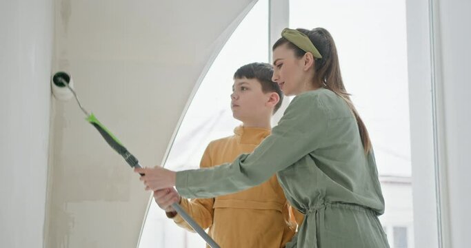 A mother and her teenage son work together. A family takes part in a home improvement project, painting a bright white wall in a new apartment. Captures the essence of DIY spirit and family bonding.