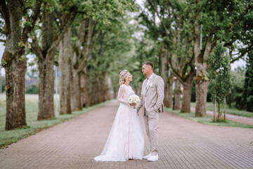 Valmiera, Latvia - August 10, 2023 - A bride and groom stand on a tree-lined path, looking at each...