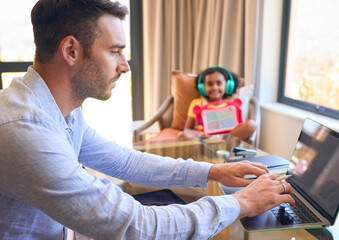 Father Working From Home On Digital Tablet As Daughter Listens To Music And Plays In Background