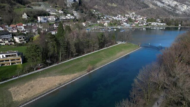 Drone clip over calm straight river at the foothills of the Swiss Alps, running past a traditional village