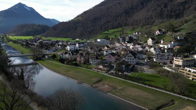 Panning drone clip of scenic Swiss village, with snowcapped mountains in the background and calmly flowing river with bridge
