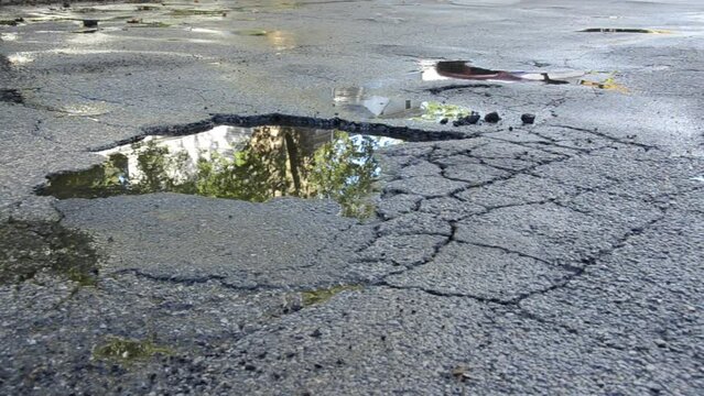 Huge pothole in street filled with water, shimmering trees and sky