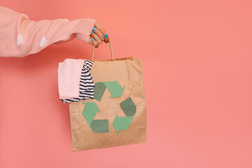 Paper bag with recycling sign with old clothes in hand against pink background.