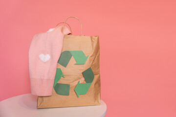 Paper bag with recycling sign with old clothes on table in front of pink background.