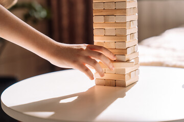 A child slides a wooden block of Jenga game on a white table under sunlight.