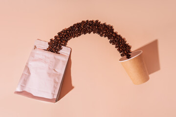 Coffee beans spill out of packaging from a paper cup on a beige background.