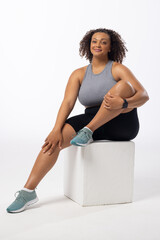 A biracial young female plus size model sits on white block, white background, copy space