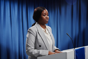 Young African American female politician in formalwear keeping her hands on platform while looking at public and speaking in microphone