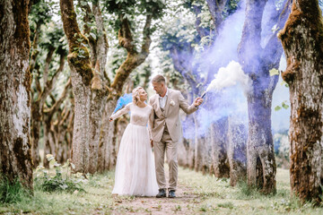 Valmiera, Latvia - August 10, 2023 - Bride and groom walking down a tree-lined path holding smoke...