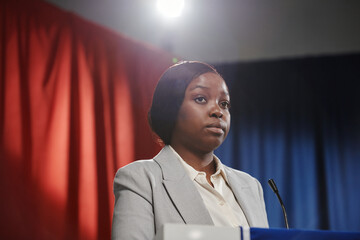 Young serious African American female delegate in formalwear standing by platform with microphone and making report for public