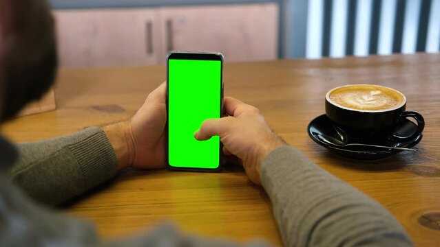 Young man sitting at cafe holding smartphone green mock-up screen in hand. Male person using chroma key mobile phone. Vertical mode. Touching, swiping display, tapping, surfing internet social media