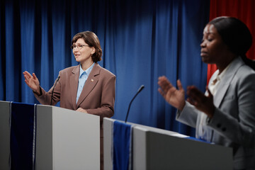 Mature brunette female speaker in formal suit gesticulating by hand and looking at public while standing by platform and making report