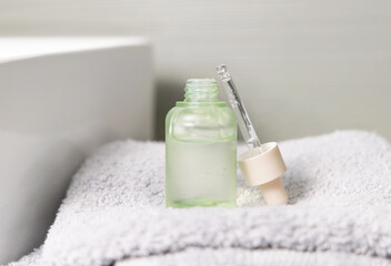 Opened cosmetic bottle with green serum on folded bath towel against basin close up