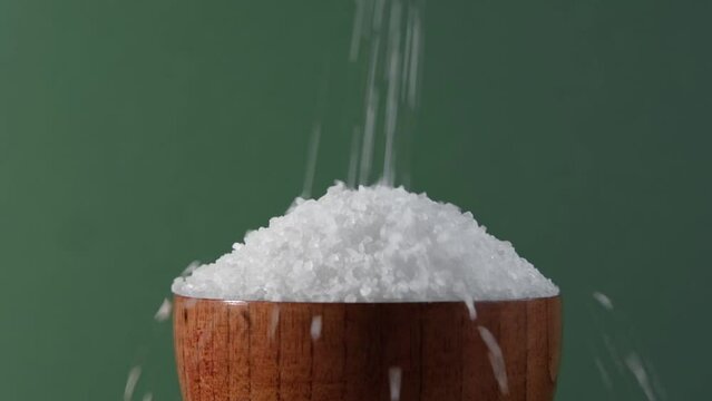 White table salt granules falling into a pile. Wood bowl. Gray color studio background. Condiment particles dropping. Clean cosmetic bath salt pouring footage. Closeup of grained sea salt pieces hill