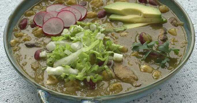 A bowl with vegan Pozole Verde dish with hominy, beans, tomatillo, and button mushrooms topped with avocado, red radish, and shredded napa cabbage. Table spin. 