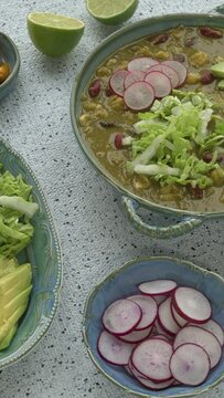 Vegan Pozole Verde dish with hominy, beans, tomatillo, and button mushrooms topped with avocado, radish, and shredded napa cabbage. More toppings on the table. Table spin. Vertical video.
