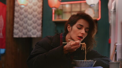 Thoughtful woman observes night city street through window at eating Chinese meal using chopsticks