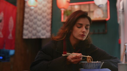 A millennial girl is eating Asian noodles with chopsticks. Woman eats street food in a Chinese cafe