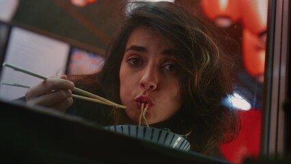 Brunette woman smiles happily eating spicy Chinese noodles with chopsticks in Asian street cafe