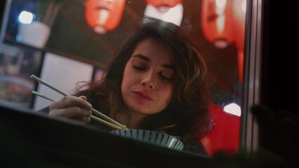 A millennial girl is eating Asian noodles with chopsticks. Woman eats street food in a Chinese cafe