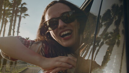Woman in sunglasses with happy smile waves hand out of car window riding along road with palm trees