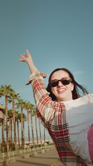 Cheerful young woman in sunglasses raises hand leaning out of car window against tall palm trees
