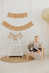 Obraz premium Baby first birthday concept. Portrait Happy caucasian little boy sits against background of holiday decorations and flags with inscription.