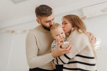 Obraz premium Happy baby boy receives kisses from his loving parents, creating heartwarming scene during first birthday celebration at home. Lifestyle moment of scandinavian family