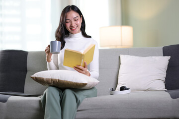 Happy young Asian woman sitting on the sofa in the living room at home reading a book and drinking...