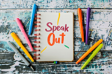 Speak Out" adorns a notebook cover - surrounded by colorful pens - urging individuals to share their bullying experiences and break the silence
